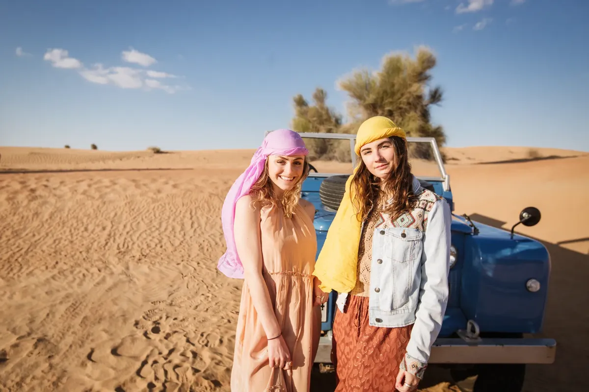 Two women smiling in the Dubai desert with a vintage Land Rover hinting at adventure and travel.